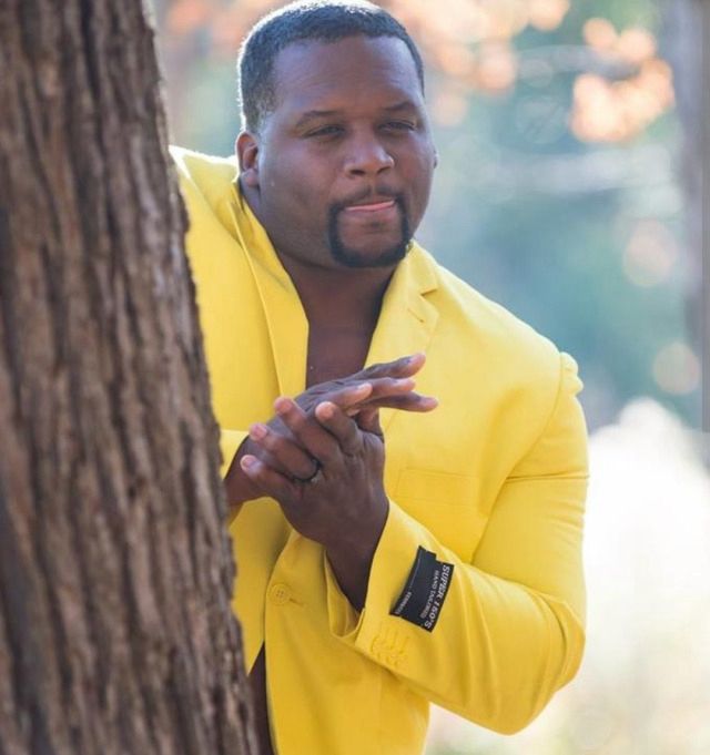 a man in a bright yellow jacket looking mischievously out from behind a tree, rubbing his hands together like a cartoon villain.