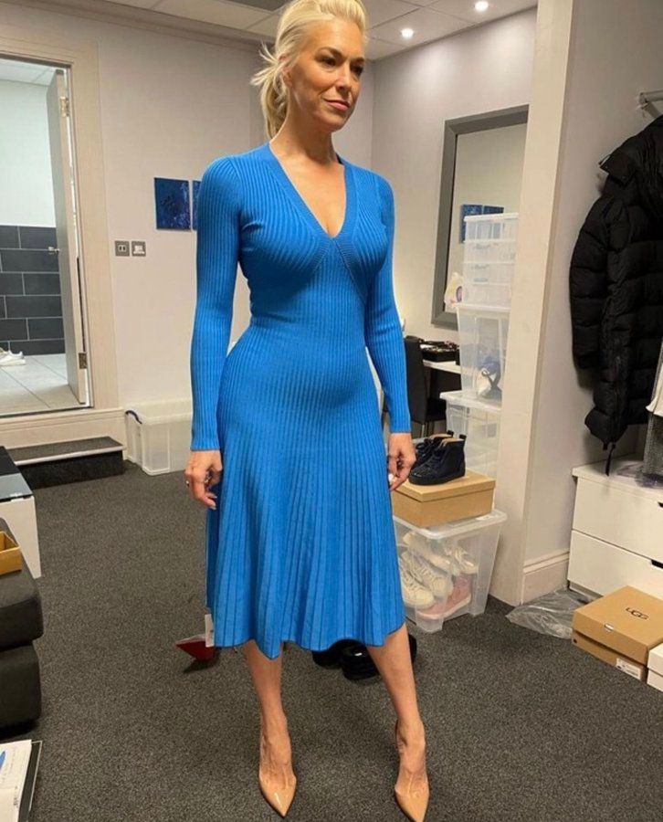 Hannah Waddingham in a sky blue, v-neck, long-sleeved, knee-length dress. The dress is form-fitting until the waist where it flows out a little bit. It fits her so well, it looks like it was made for her specifically. Photo made for a costume check for Rebecca Welton, for Ted Lasso season 3, posted by head of costume Jacky Levy (at some point).