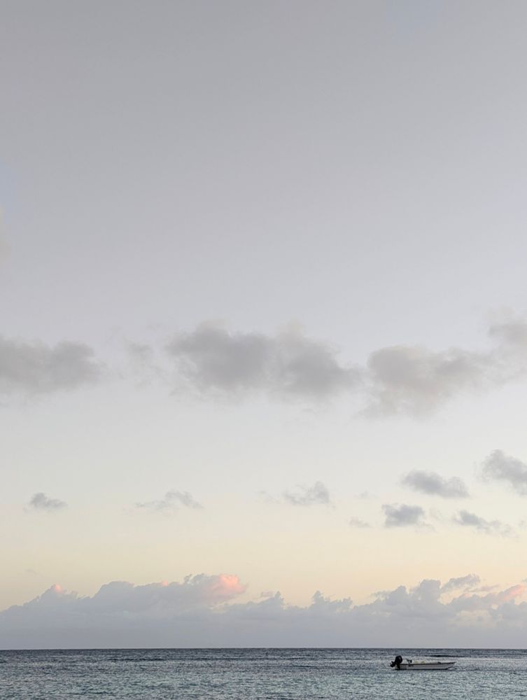 Image of a sunset, the sky is pale grey, the clouds are dark grey & dusty rose.
A boat can be seen floating on the water.