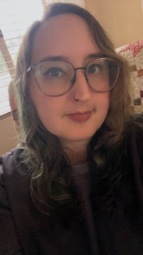 selfie of me with a black sweater and large glasses, my hair is brown/greenish and goes just below my shoulders, i have silver and black eye makeup and dark pink lipstick, i'm smiling slightly 