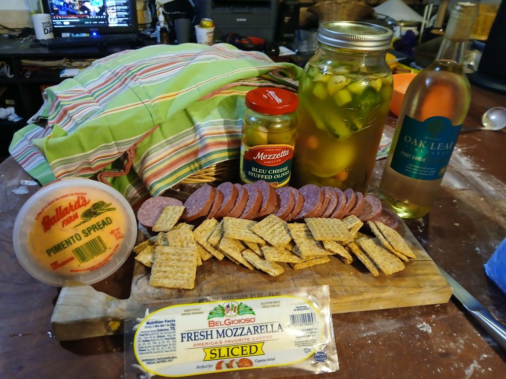 A charceuterie board, from left to right, a pimento cheese spread, summer sausage rounds, in front of that are olive oil and rosemary triscuit crackers, and in front of that, a log of mozzarella. Behind the board is a basket of bread covered with a green striped cloth, a jar of bleu cheese stuffed olives, a jar of pickled vegetables, and a bottle of extremely cheap, and strong, pinot grigio wine. 
