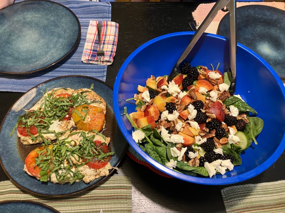 Caprese chicken with fresh basil, mozzarella, and heirloom tomatoes drizzled with balsamic on a blue plate, beside a big blue salad bowl with spinach, cucumbers, peaches, blackberries, pecans, and fresh mozzarella.