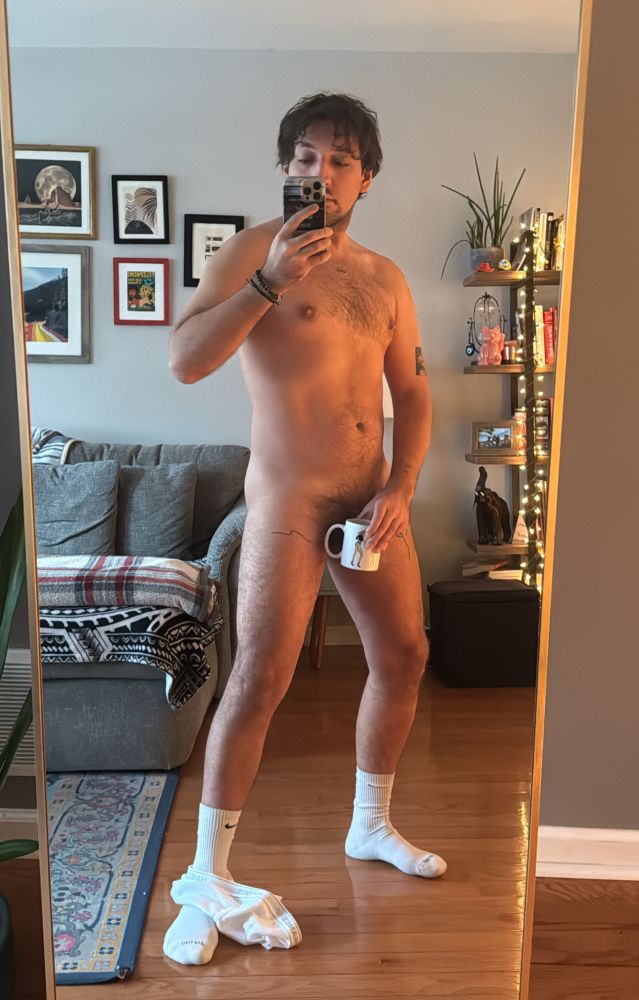 Nude in floor mirror with reflective holiday lighting and using a mug to cover peepee 