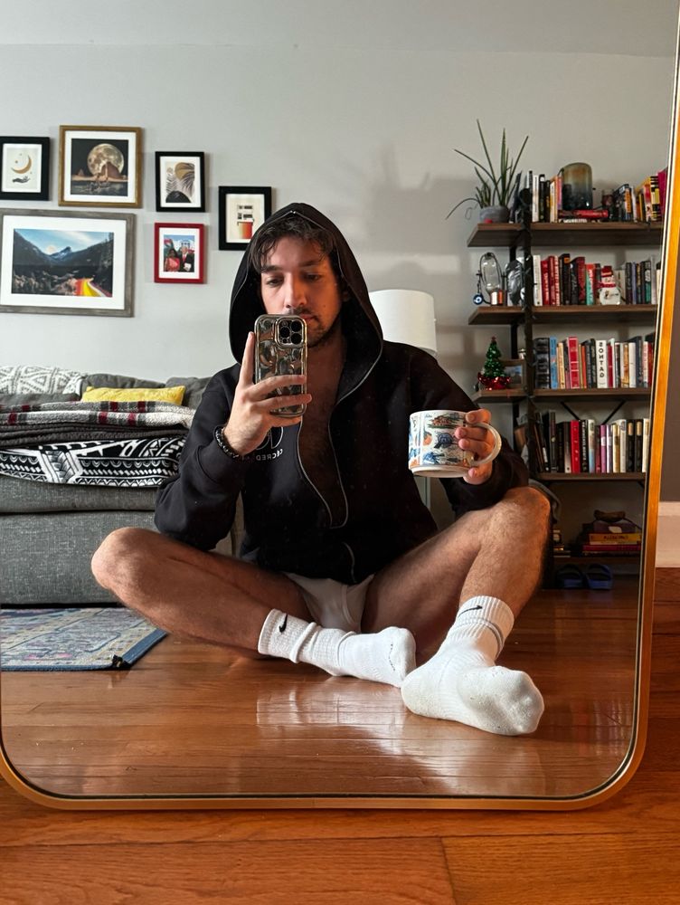 sitting on the floor looking in the mirror nearly cross legged holding mug of coffee and taking a photo in a zip hoodie and no pants 