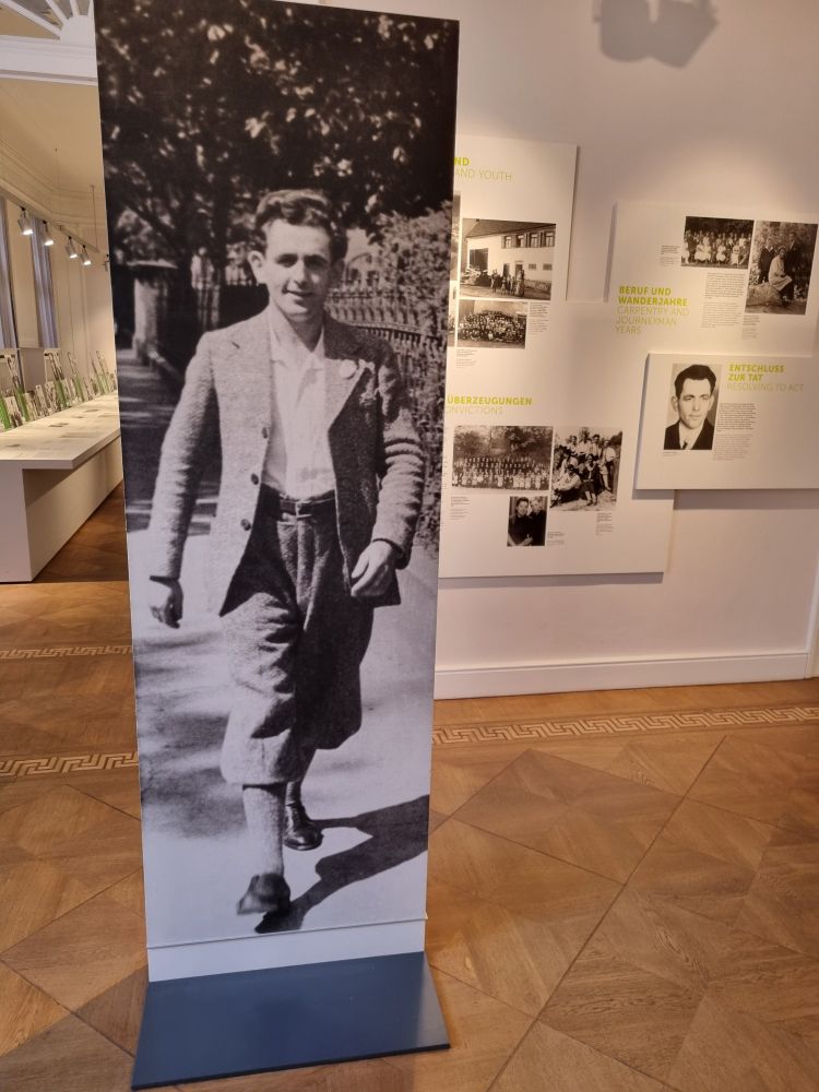 Aufnahme mit Portrait Georg Elsers, im Hintergrund ist ein Teil der Ausstellung zu sehen.