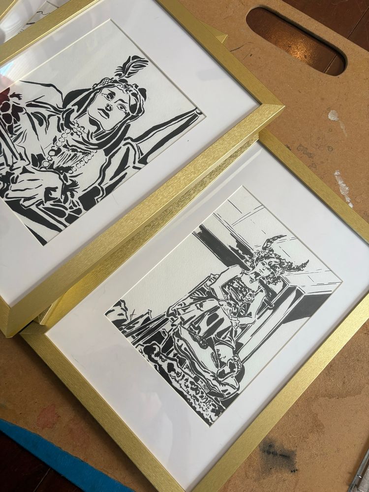 Two ink drawings of women in a gold frame on a well loved drawing board