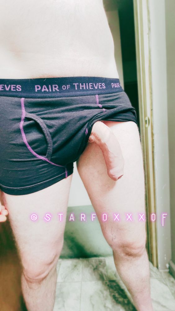 Starfoxxx stands in a doorway wearing black and pink boxers. One side is tucked up and his erect penis hangs out of them. 