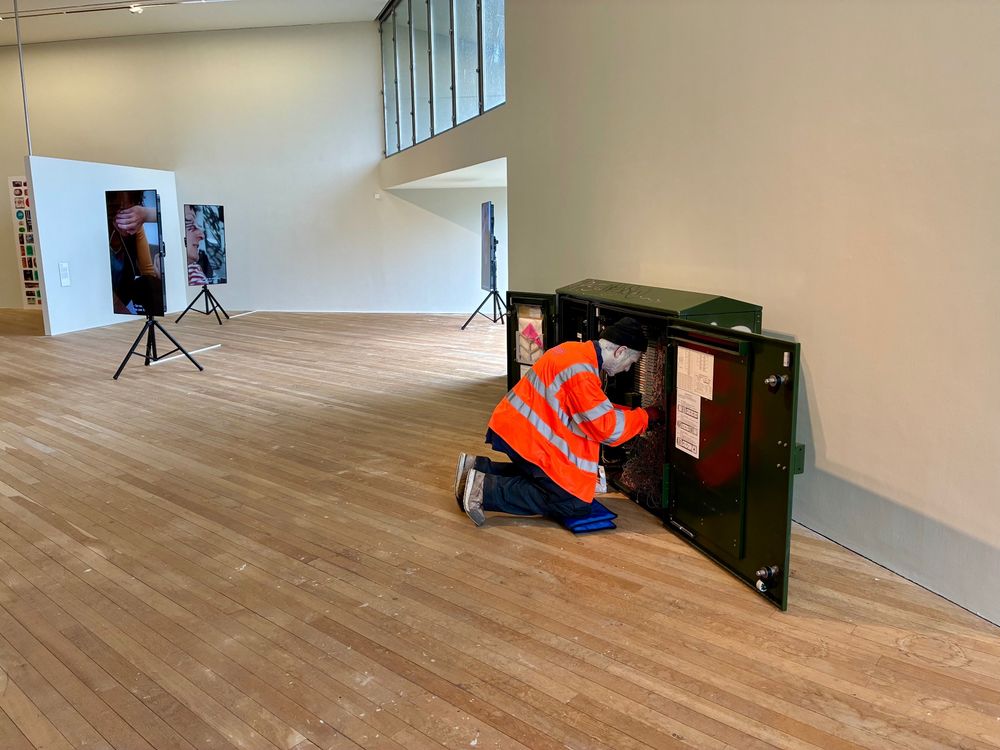 A realistic sculpture of a male engineer, wearing a hi-vis jacket, kneeling in front of a communications box, ostensibly repairing it. The setting is an art museum.