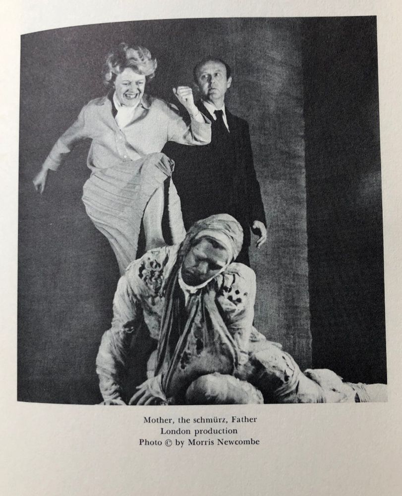 Production photo of Boris Vian's play The Empire Builders. Image shows a woman ('Mother') vigorously kicking a bandaged man ('the schmürz') who is on the floor, while another man ('Father') looks on.