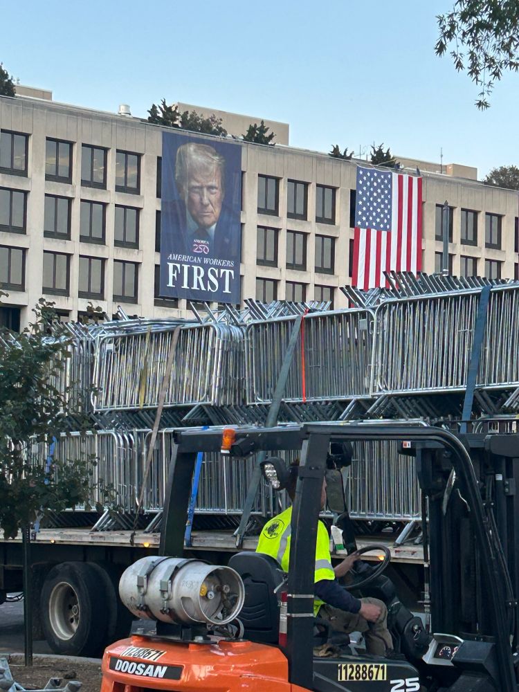 Crowd control barricades on the back of a truck. In the background, a government building with a massive Trump banner. 