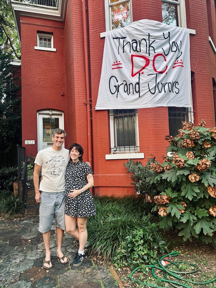 Nerdy couple standing in front of a house with a banner reading “Thank you, DC grand jurors” and two little DC flags. 