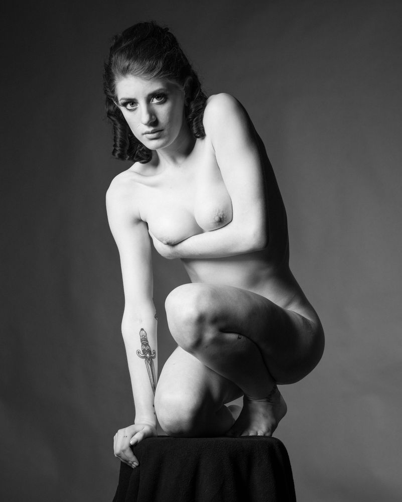 A nude woman cradling her breasts with one arm crouches on a plinth, and fixes us with a subtly challenging stare. She is lit from above-left and the background is an irregular grey gradient.