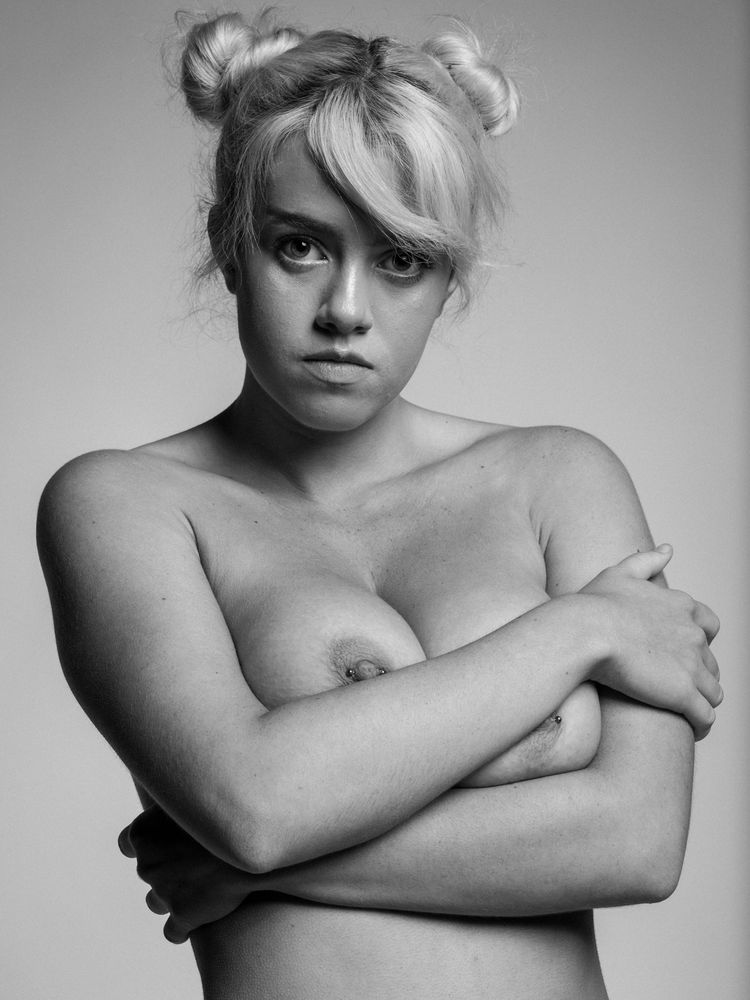 Head and shoulders image of a nude woman with blonde hair done up in two space buns and her arms crossed in front of her chest.