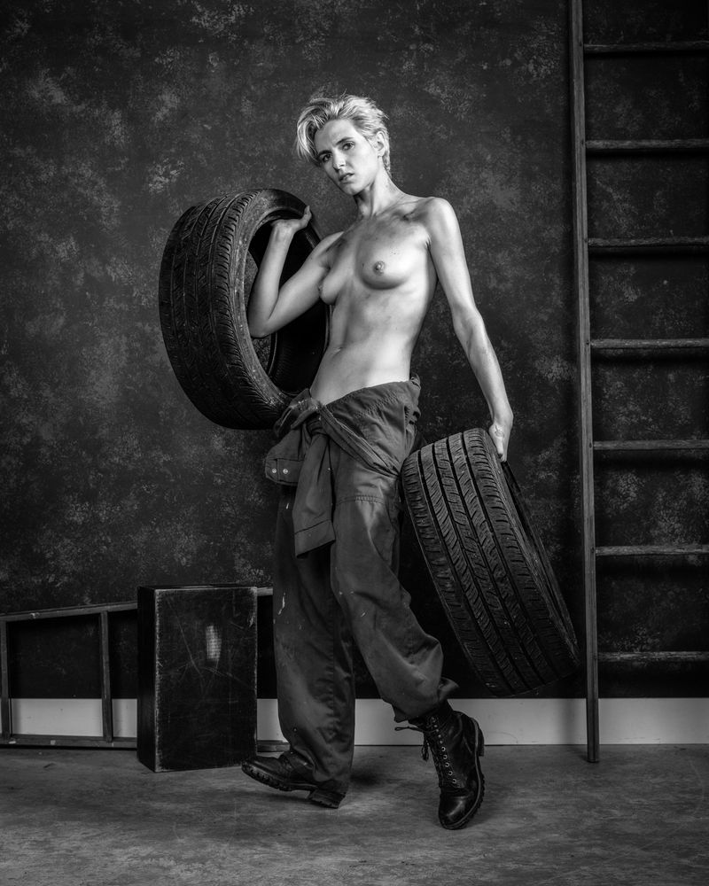 A woman with short hair, her skin covered in grease marks and dirt, wearing coveralls with the top tied at her waist is hoisting two fat performance auto tires. There are two ladders visible behind her, leaning against a mottled wall.