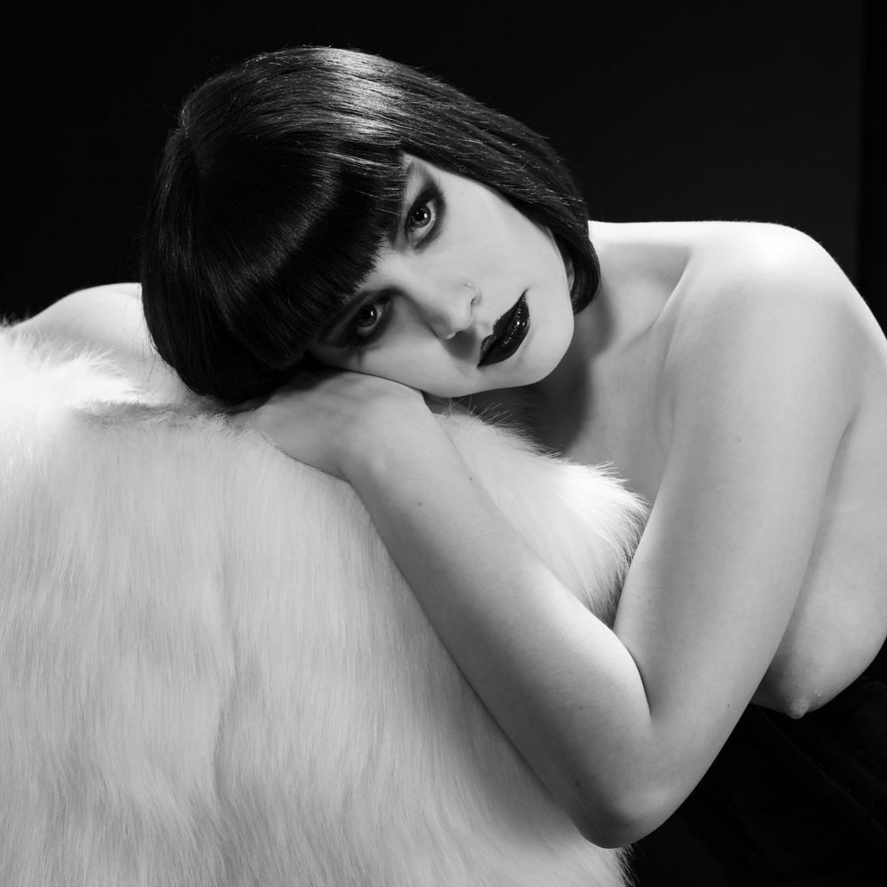 A woman with her hair in a bob with bangs and glam makeup leans on a faux fur throw. She's also topless. She's lit with high contrast lighting.