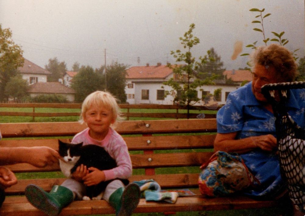 Klein-Katrin (rosa Pullover, graue Jogginghose und grüne Gummistiefel) sitzt mit ihrer Oma (blaue Kittelschürze) auf einer Bank irgendwo in Bayern, vermutlich in Wertach. 

Im Arm hält Klein-Katrin eine große schwarzweiße Katze und guckt dabei glücklich in die Kamera. Von der linken Seite streichelt jemand (Opa oder Onkel?) die Katze. 

Insider: Ich hatte damals schon, wie ich im Nachhinein erfahren, habe eine Katzenallergie und bekam damals wahnsinnig schlecht Luft. Ich ließ die bayrische Kuschel-Katze trotzdem nicht los! 