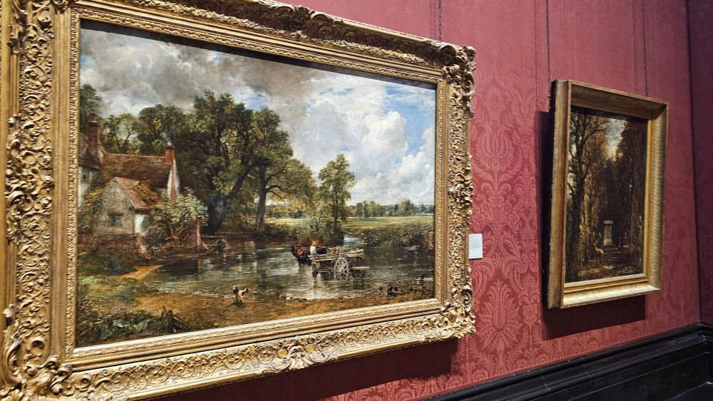 A photograph inside of The National Gallery displaying the two aforementioned paintings. The wallpaper is red and both paintings are hung in elaborate gold frames.