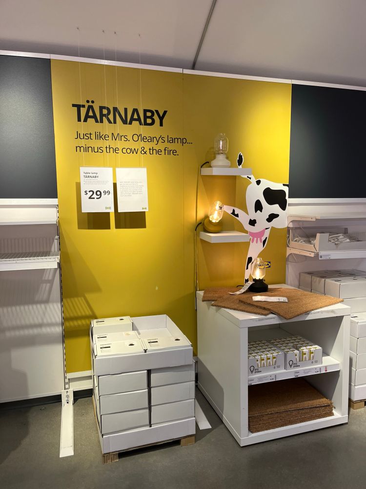 Photo of a lamp display at IKEA. A cow is kicking over a lamp, and text above reads “Just like Mrs. O’Leary’s lamp…minus the cow and the fire”