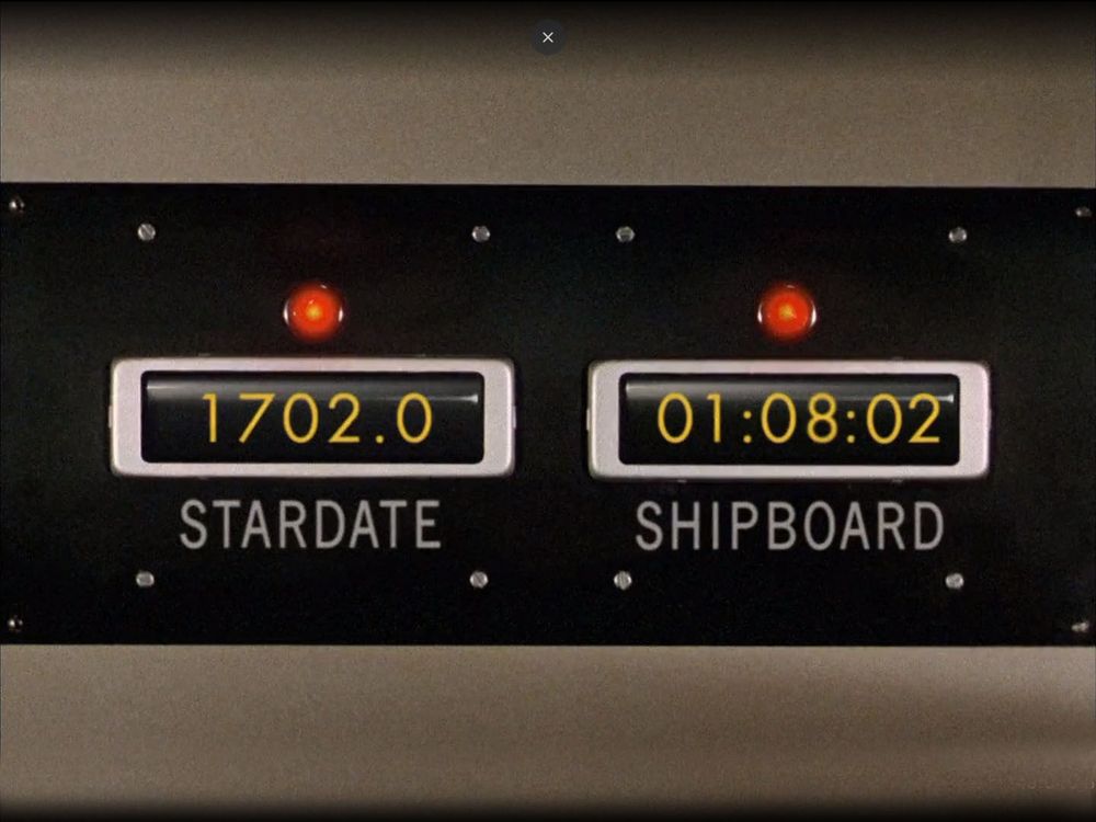 a long black metal panel screwed into beige plastic, with two numerical displays in white frames, above each of which is a large round red light, lit. They read STARDATE 1702.0 - SHIPBOARD 01:08:02