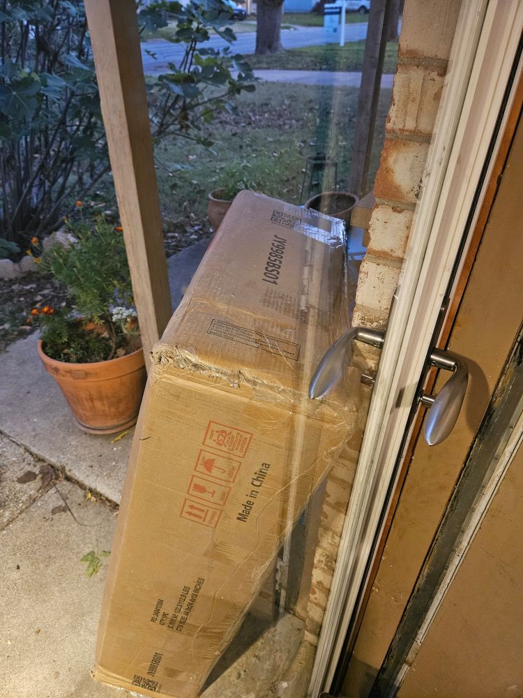 Picture is taken through a glass door. A very large box on the porch is propped against the handle, leaving essentially 2 options: heave the door open, knocking the box out of the way & breaking the contents, or wait for someone to come to the door to assist. 
Potted marigolds and a fig tree are visible in the background. 