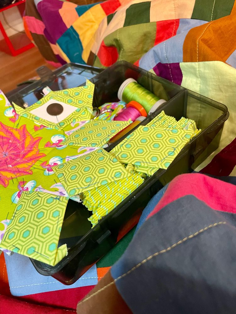 green epp piece sitting on top of a plastic box filled with thread basted epp shapes and thread spools on top of a multicolor bar quilt 