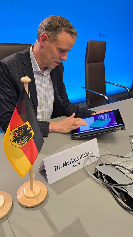 Dr. Markus Richter sitzt am Tisch der Pressekonferenz und schaut auf ein Tablet mit Anzeige der Studie eGovernment Monitor 2024 der Initiative D21. Im Vordergrund die Bundesfahne an einem Holzstempel. 