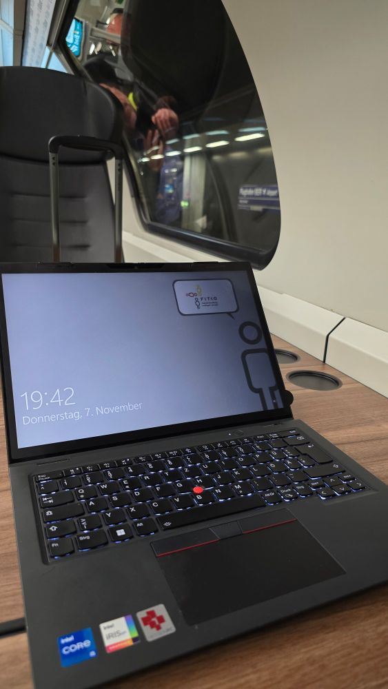 Laptop auf einem Tisch in der Regionalbahn mit Sperrbildschirm und der Anzeige #FITKO und Uhrzeit 19:42. 