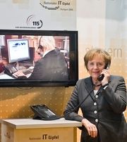 Alt-Budeskanzlerin Merkel telefoniert auf dem Kongress IT-Gipfel 2009 symbolisch mit der einheitlichen Behörden-Hotline Nummer 115. Sie hält ein schnurgebundenen Telefonhörer am Ohr, dahinter ein Bildschirm mit dem Bild eines Call-Center Mitarbeiters. 
