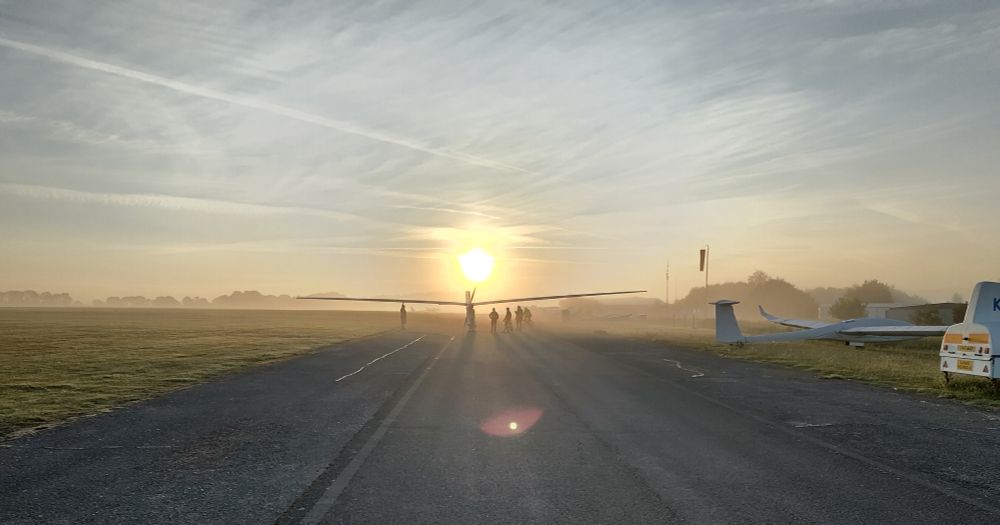 A group of people walk a slender aircraft with long wings down a tarmac taxiway, all silhouetted by the rising sun ahead of them. A shallow mist rises off of the airfield's grass surface.