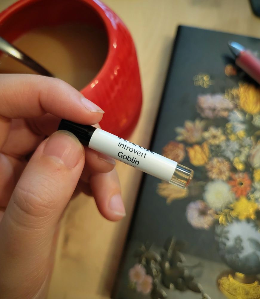 Hand holding a perfume sample by Poesie called "Introverted Goblin"
In the background there's a mug of tea and a journal + pen 