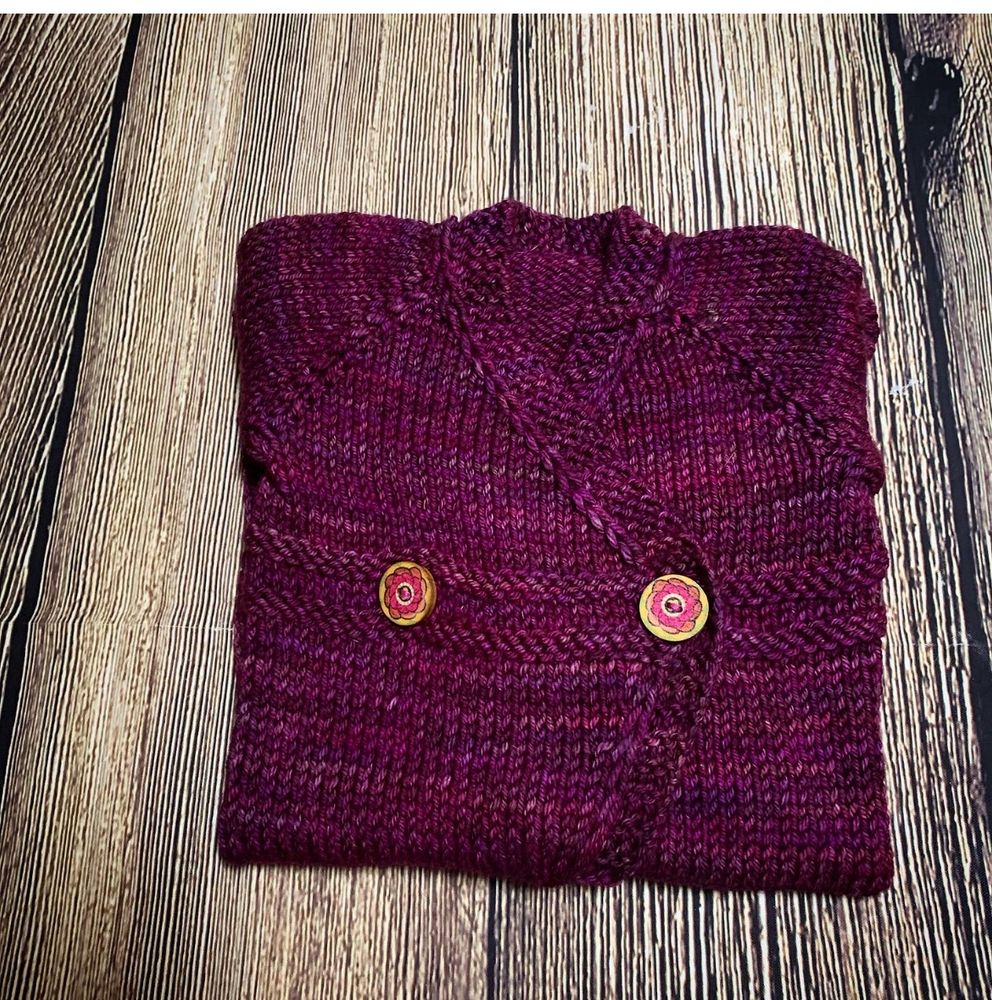 Carina Spencer’s Small Things Romper, folded up for wrapping. This version is knit in a deep pink toned purple. It has wooden buttons with pink flowers pintes on it.