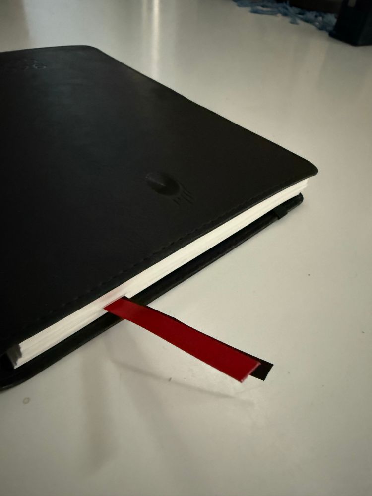 Schwarzer Planer mit weißem Papier und herausschauenden roten und schwarzen Lesebändchen auf weißem Tisch.