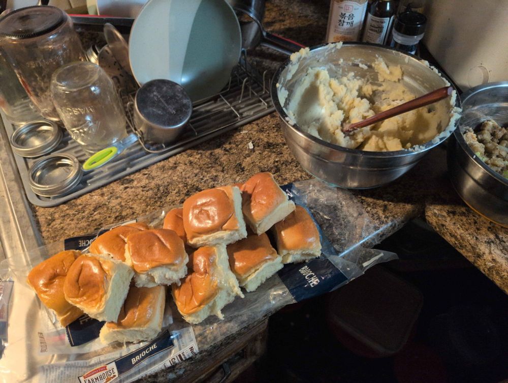 a pile of small rolls, a metal bowl of smooth mashed potatoes and.. half of a metal bowl filled with bread stuffing