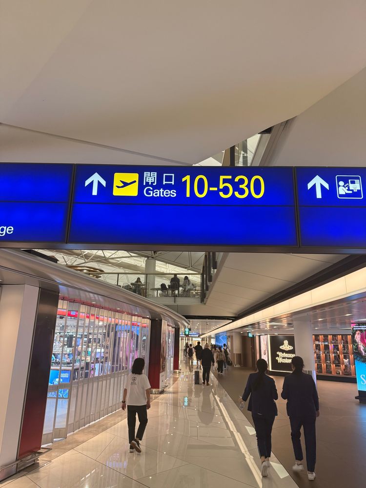 Alt text: A blue and yellow airport sign overhead directs travelers to gates 10–530, with arrows pointing straight ahead. The corridor below has a shiny tiled floor, people walking in both directions, and shops lining the sides, including a storefront with red lights on the left and another labeled “Victoria’s Secret” on the right. An upper level with seating and people is visible above.