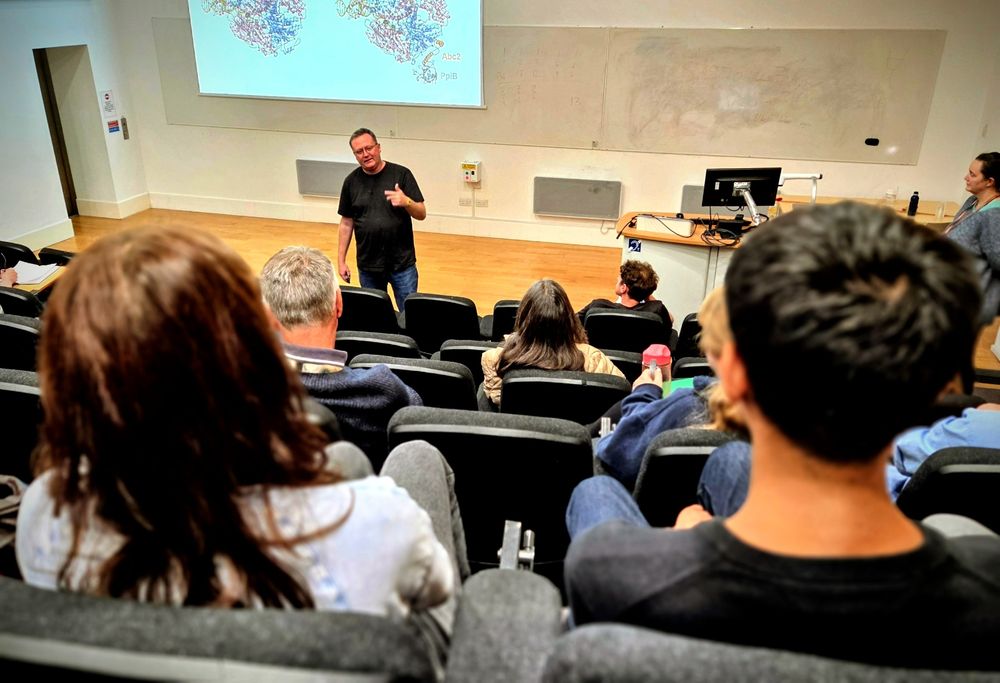 Mark Dillingham facing the audience while presenting a talk at the University of York. Protein structures of the RecBCD complex are shown on the projector screen. 