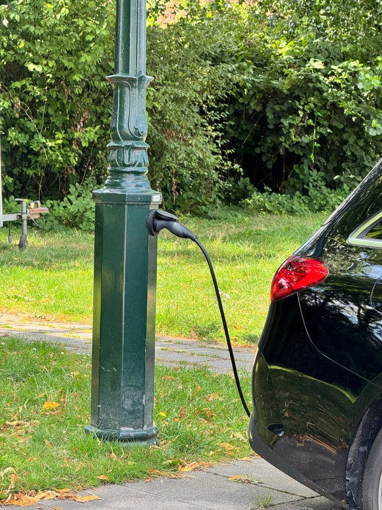 Eine altertümliche grüne Straßenlaterne, davor ein E-Auto, dazwischen ein Ladekabel, dass an einer Ladesteckdose am Laternenmast angeschlossen ist.
