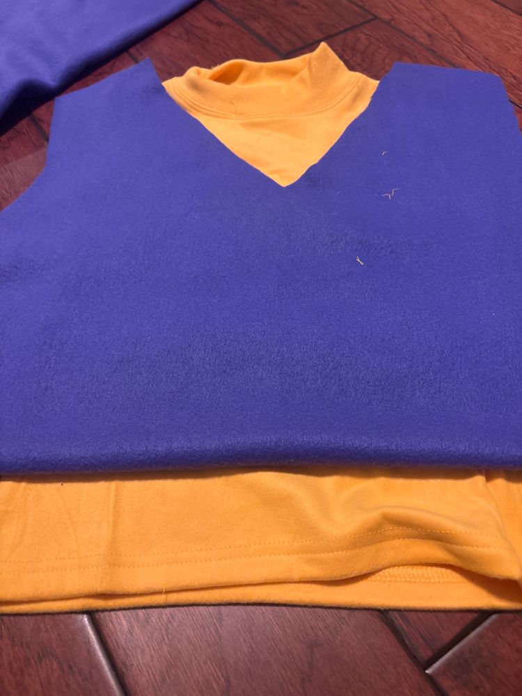 A purple front shirt pattern resting on top of a  golden sleeveless turtleneck. Both are on a cherry wood floor. 