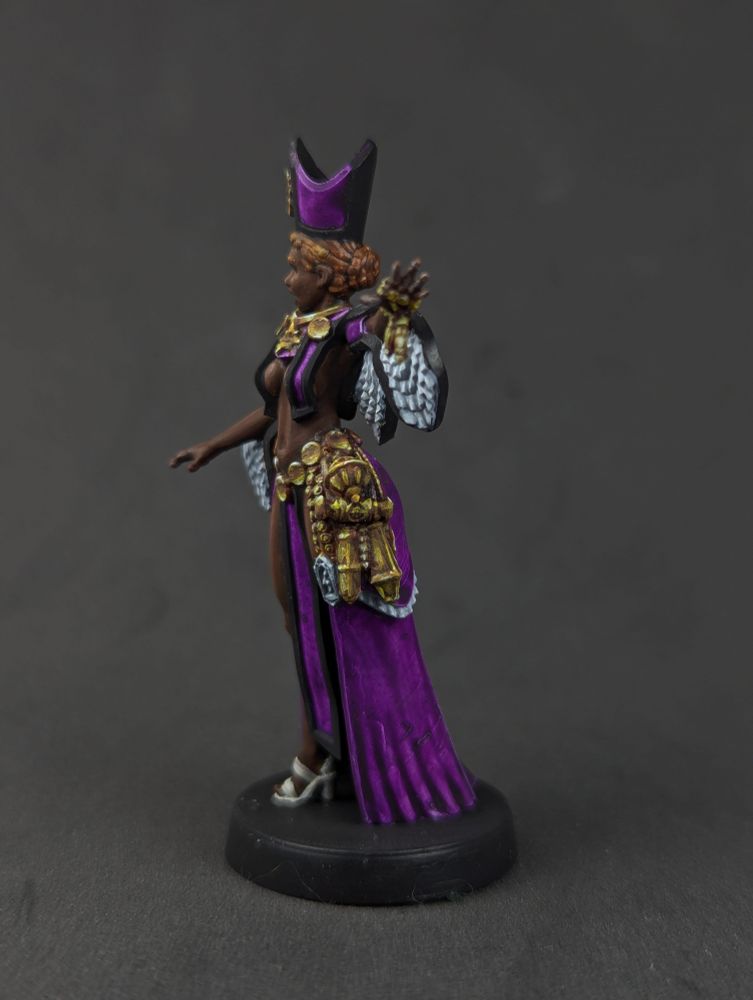A black priestess in purple robes with gold jewellery. From the side showing more of the jewellery details.