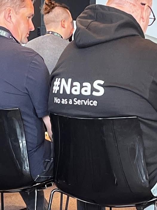 The photo is of the backs of people sitting in black plastic chairs, like at a conference. The person on the left is only half seen and wears a dark blue polo. The person the image is focused on wears a black hoodie with white writing on the back that reads “#NaaS
No as a Service”