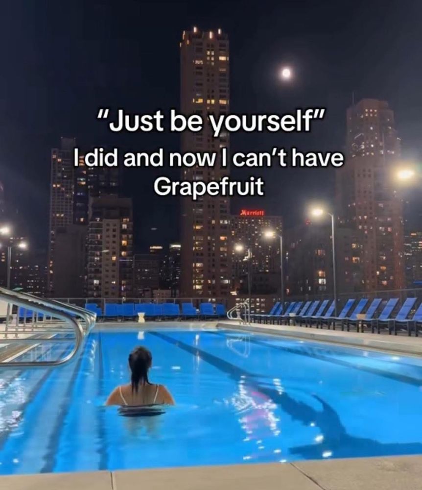 A photo of a person with shoulder length hair facing away from us and standing almost shoulder deep in a swimming pool that appears to be on the top of a building. Blue chairs surround the pool on the deck and the person looks out onto a city skyline at night. The overlaid meme text says “Just be yourself" I did and now I can't have
Grapefruit.”