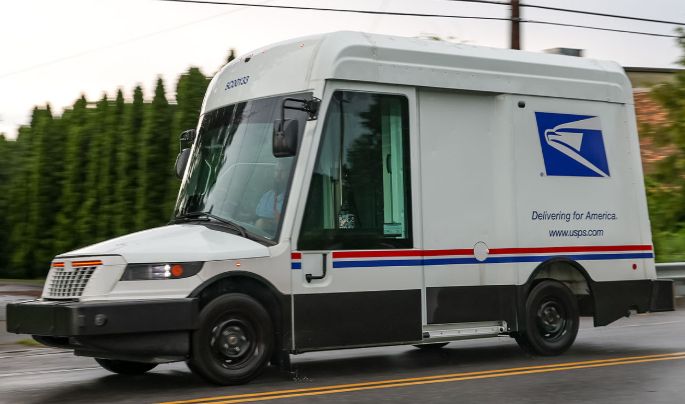 The new electric mail truck, made by Gruman, driving to the left of the image but taking up the full frame from a 3/4 view. It has a large windscreen, which combined with small lights, an outstretched hood, and a grill with a large wide bumper taking up the entire space below it, which all add up to a friendly-looking "face."