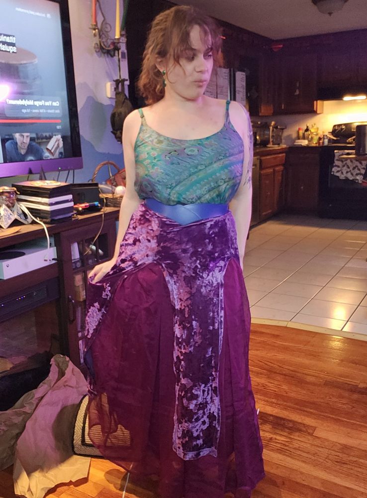 A curvy figured white person wearing a paisley teal patterned tank top and a purple velvet skirt with different textures.