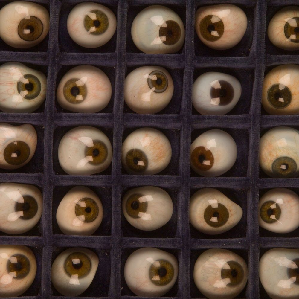 Glass eyes lying in a grid in a blue velvet case. The eyes are half globes that closely resemble a human eye, including the black pupil, coloured iris (in shades of blue, green and brown) and the white sclera with the details of red blood vessels. 
