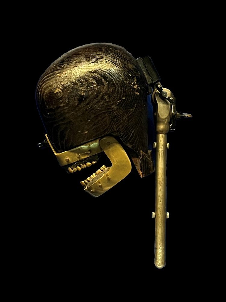 Wooden head shaped object with brass jaws and teeth mounted on a metal stand