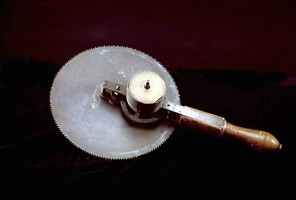 Surgical instrument consisting of a round, toothed blade attached to a wooden handle. The blade is made of a rusted metal.
