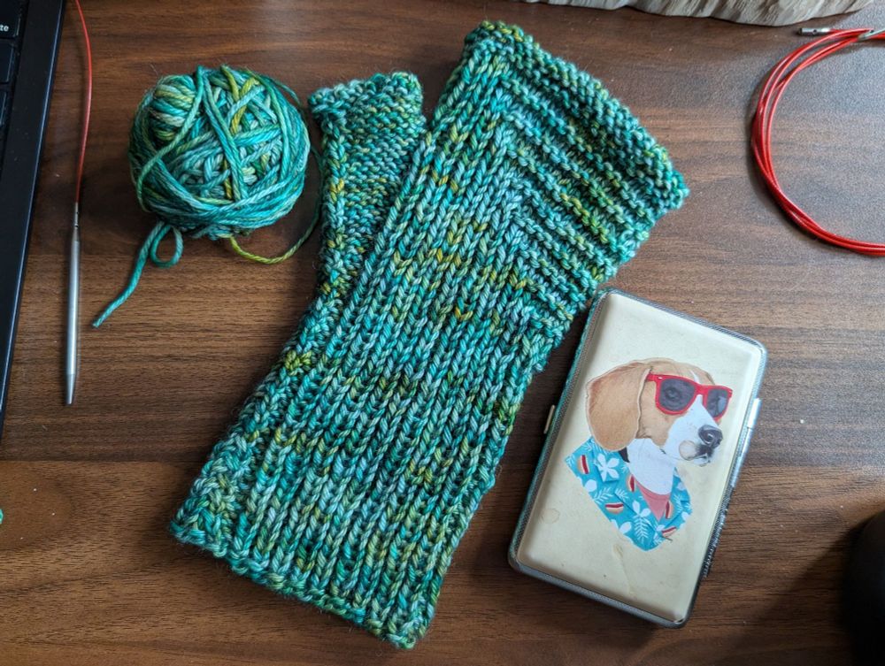 A flat lay showcasing a single fingerless glove/gauntlet-style mitt, knit in teal and olive variegated hand-dyed yarn that reminds me of sea glass. Surrounding the mitt are the leftover yarn ball, steel knitting needle on a red cable, and a notions kit with an illustration of a beagle wearing red sunglasses and an aloha shirt.