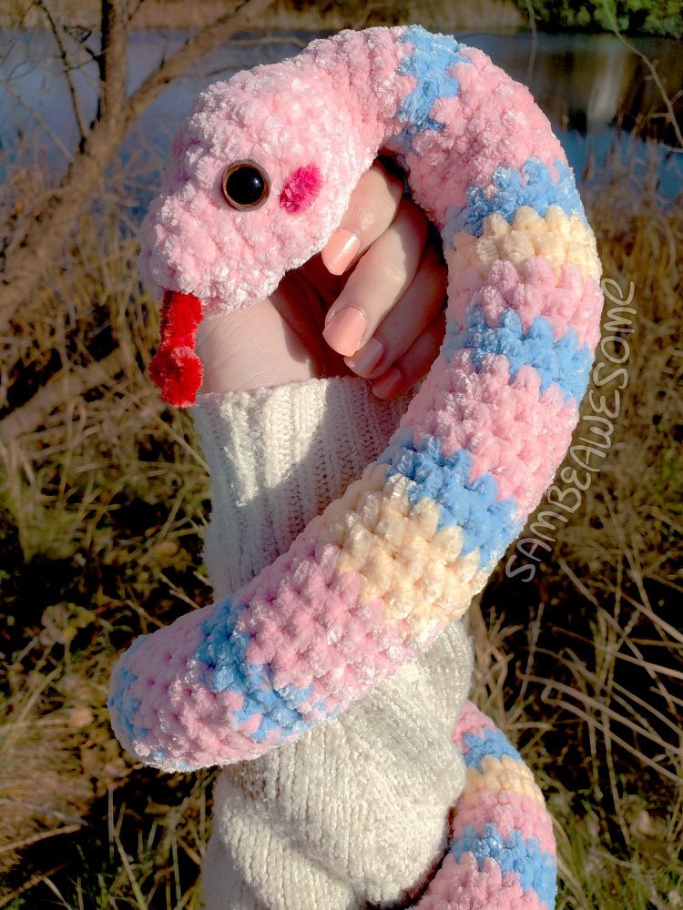 A real photo of a white person's hand holding up a crochet snake (light pink with blue and creme stripes, with a red tongue and blush, and brown eyes) in front of a nature background with bushes, a tree, branches, and a pond. The person is wearing an off-white knit sweater with light pale pink nail polish.