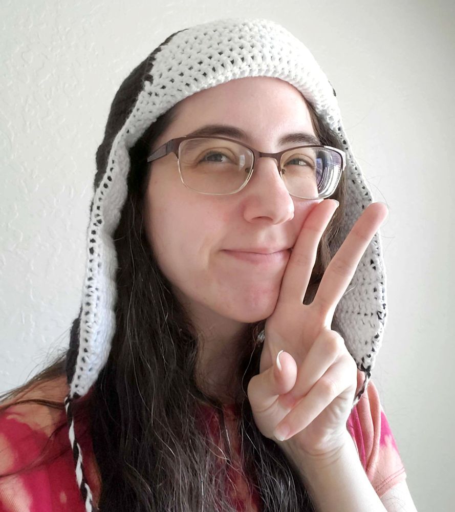 A selfie of the OP, white femme person with long dark brown hair and glasses, holding up a peace sign and smiling, wearing the crochet ushanka from the pattern.