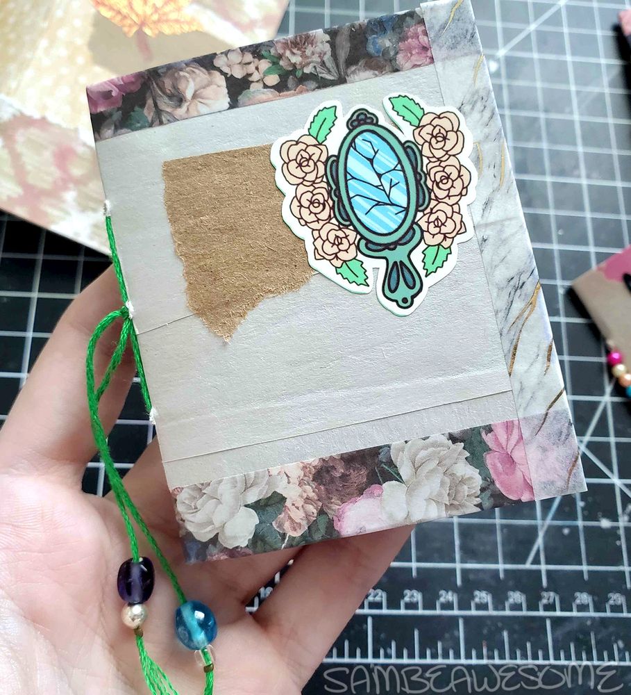 Photo of a small junk journal with off-white craft paper on the cover and washi tapes (flowers on the top and bottom, and white marble on the ends) on the edges. The cover also has torn brown packaging paper with a bad luck broken mirror sticker with flowers by Amanda Elise (Creative & Caffeinated). Hanging off the excess strands of the hand-sewn spine is various color coordinating beads.