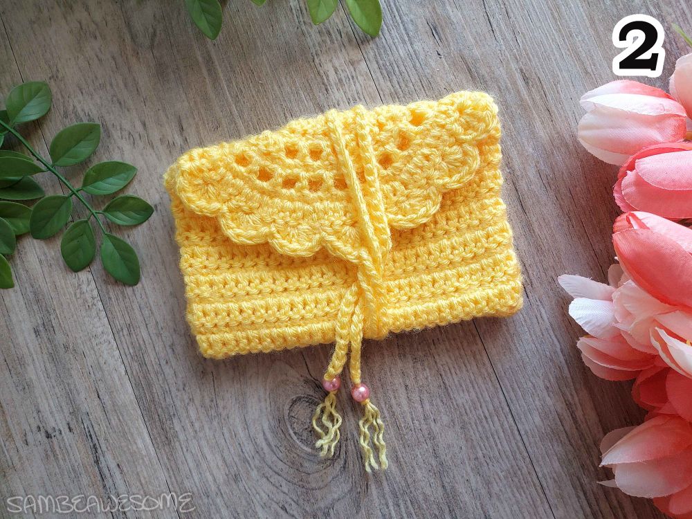 Photo of a handmade crochet pouch with a semi-circle scalloped edge flap, and a yarn chain wrap to close it, with beads on the end of the chain. The color of the bag is golden yellow with pink beads.